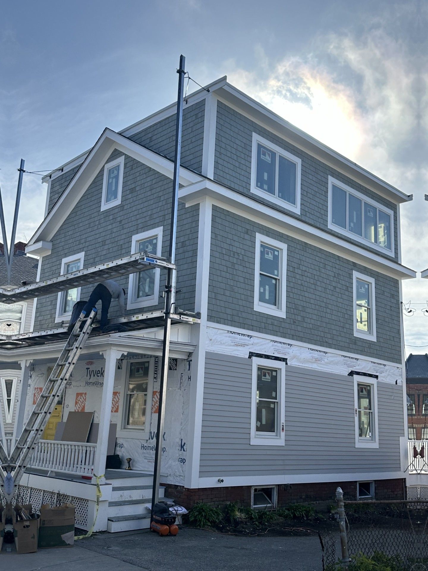 Multi-story house under construction, with gray siding, white trim, and a person on a ladder working on the exterior.