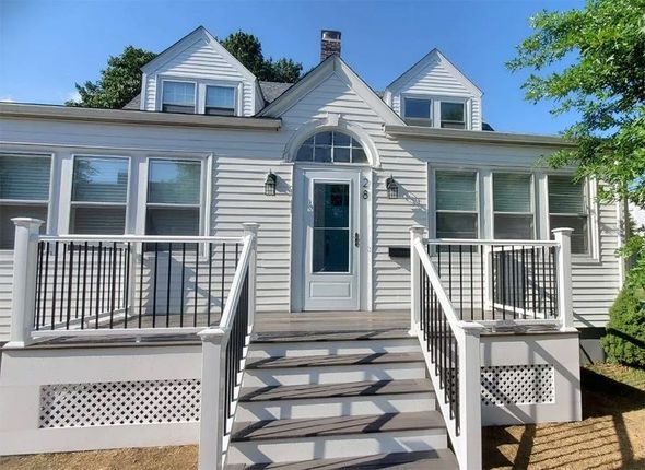 White house with porch, steps, and black railing. Dormers on roof, sunny day.