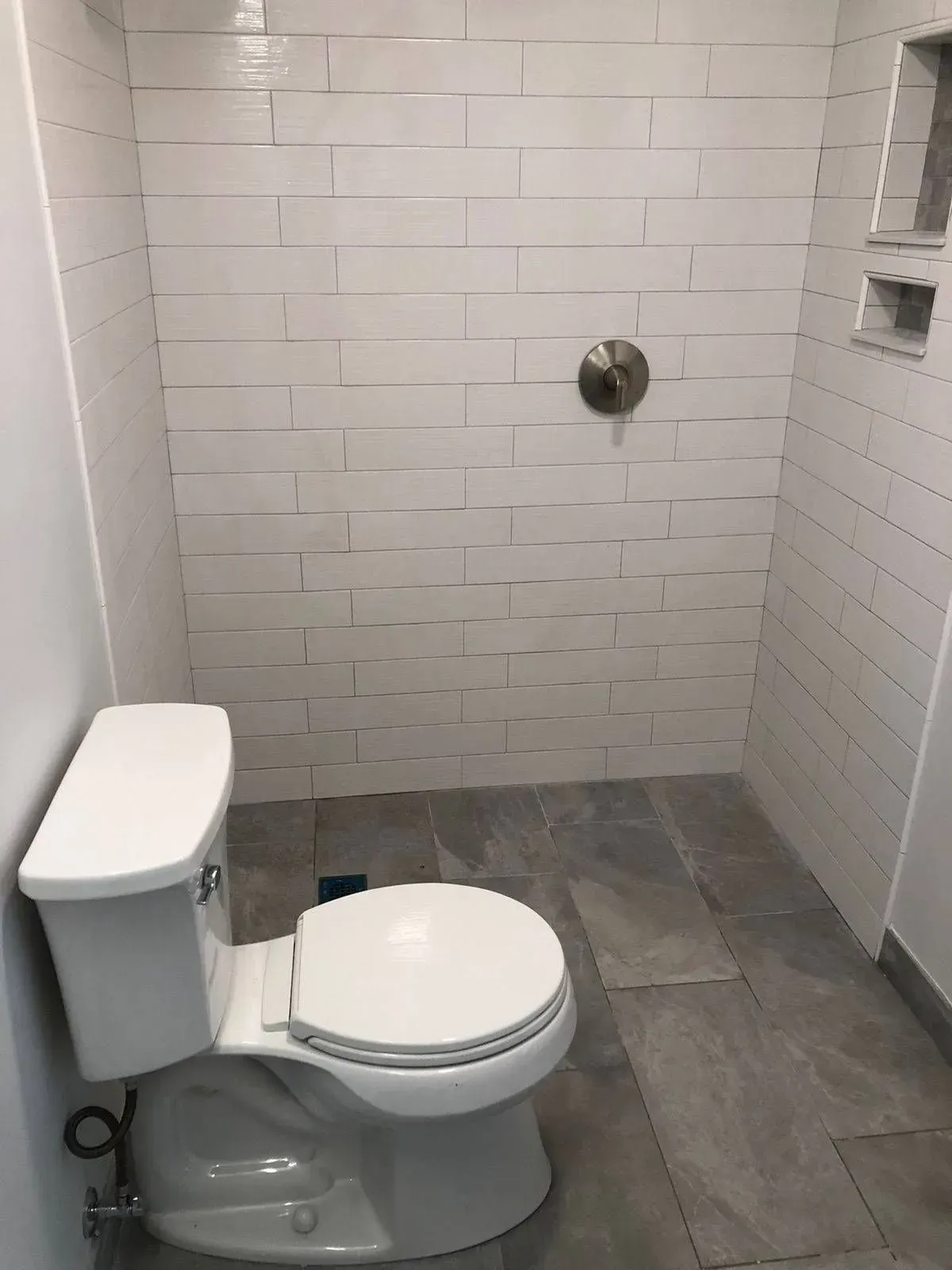 A small bathroom with a white toilet, gray floor tiles, and white tiled shower area with a shower head.