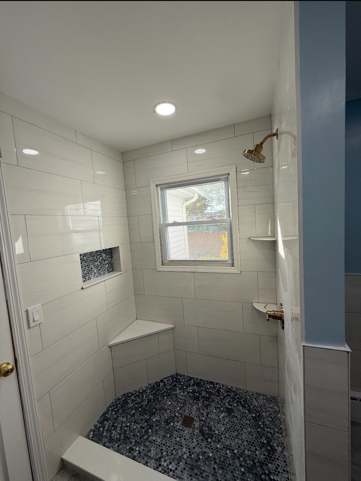 Bathroom with white tile walls, blue pebble floor, showerhead, corner seat, and window.