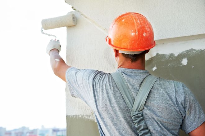 Worker in orange hard hat painting a building wall with a roller.
