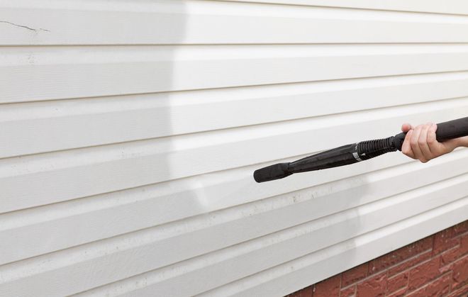 Person pressure-washing white vinyl siding beside a brick foundation.