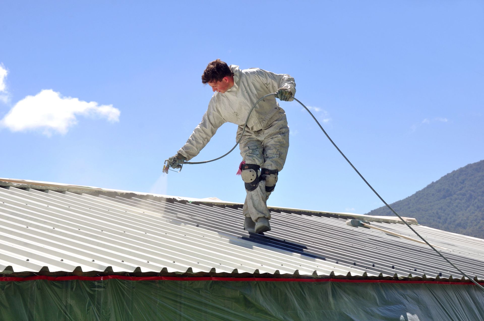 Worker cleaning a corrugated metal roof with a rope under a blue sky