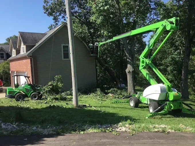 Yellow loader clearing storm debris while a worker in orange clears branches by the roadside.
