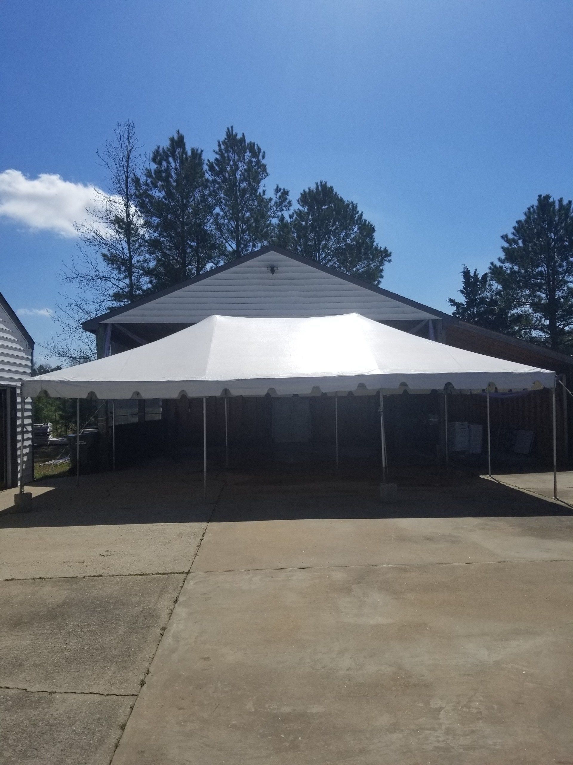 White tent set up in a concrete area, with a house and trees in the background under a blue sky.