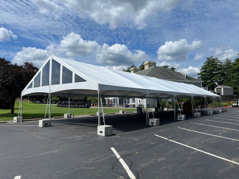 White tent set up on asphalt parking lot, next to green grass and trees, under a cloudy blue sky.