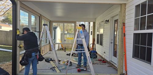 Workers renovating a covered porch, with ladders, tools, and exposed framing along the walls.