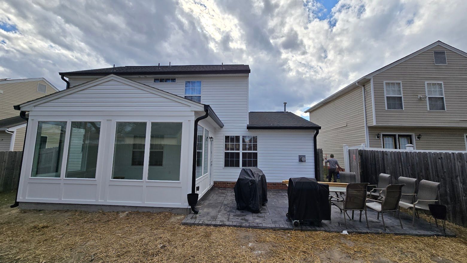 Backyard patio with white house extension, large windows, black grill covers, and cloudy sky
