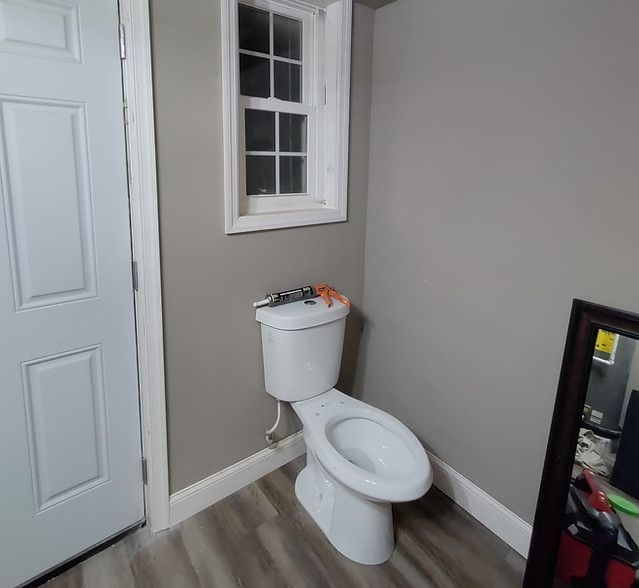 Small bathroom with a white toilet beneath a window, gray walls, and wood-look flooring.
