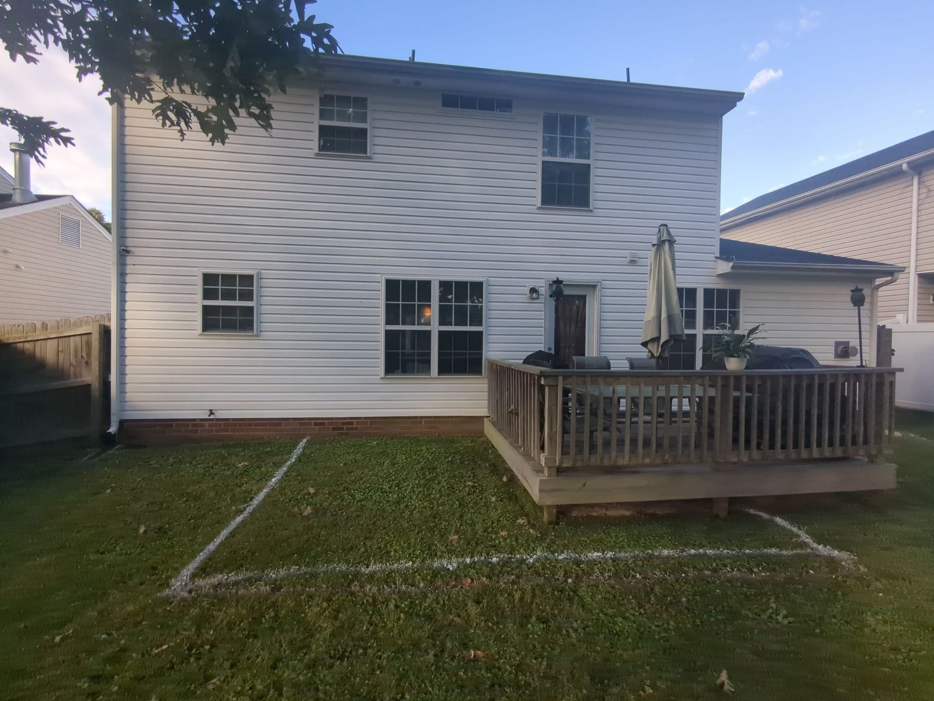 Backyard view of a two-story white house with a wooden deck and patio umbrella, surrounded by grass.