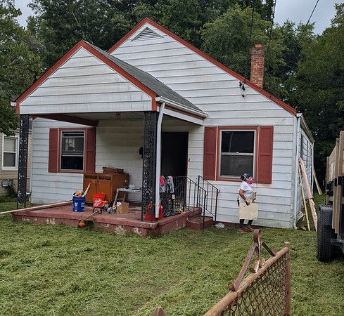 Small white house with red trim and a front porch, cluttered yard, and a child standing by the right side.
