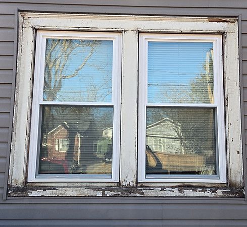 Old white-framed double window with peeling paint reflecting trees and houses.