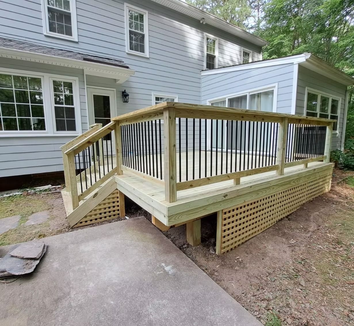 Wooden accessibility ramp with railings beside a white house, leading from a concrete path to a deck.