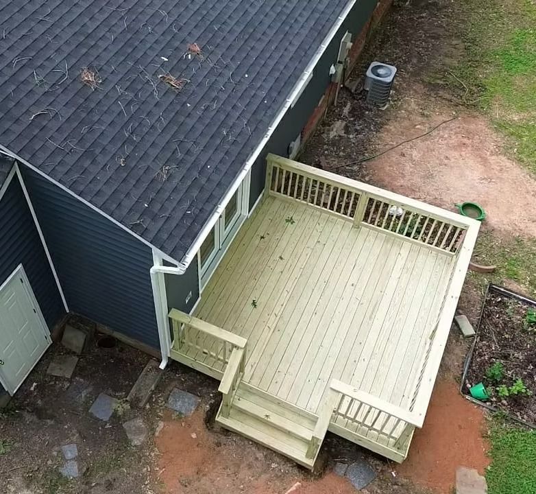 Aerial view of a dark house with a light wooden deck and steps in a muddy yard.