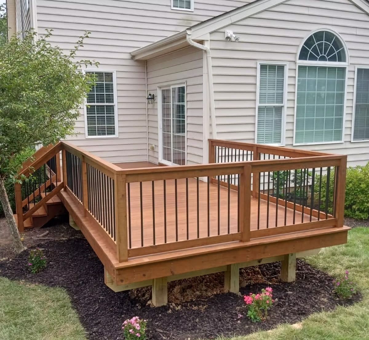 Wooden backyard deck with railings attached to a beige house, overlooking a small garden.