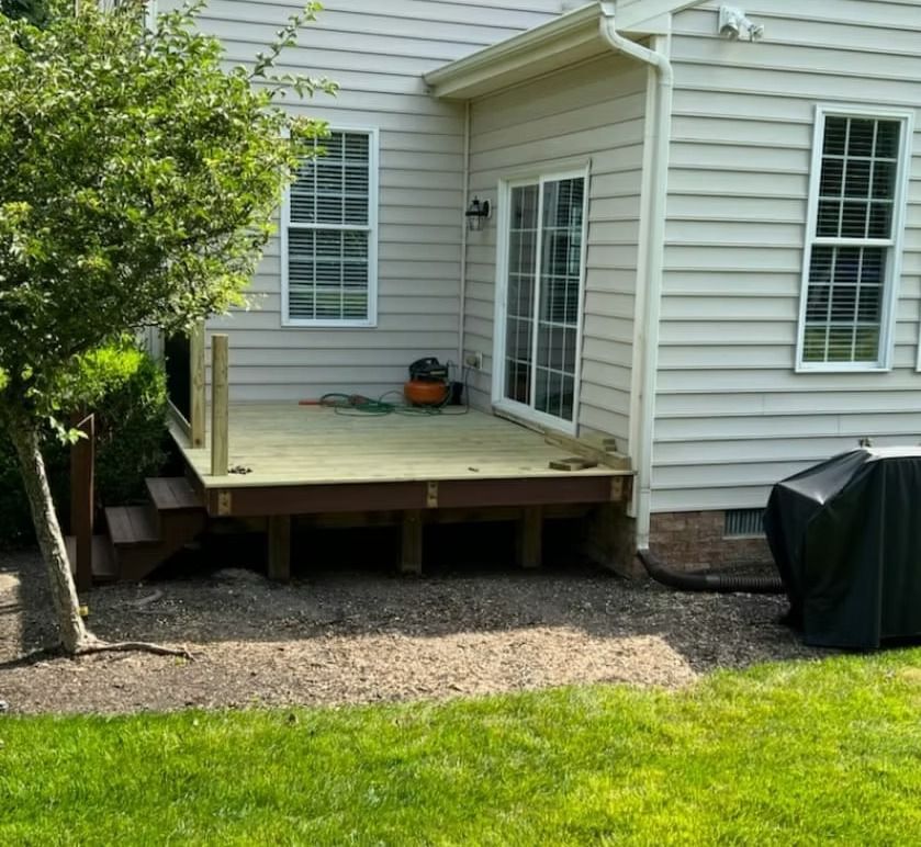 Wooden backyard deck beside a white house, with sliding glass doors and a covered grill nearby.