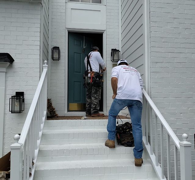 Two workers carrying tools up white front steps to a dark green front door