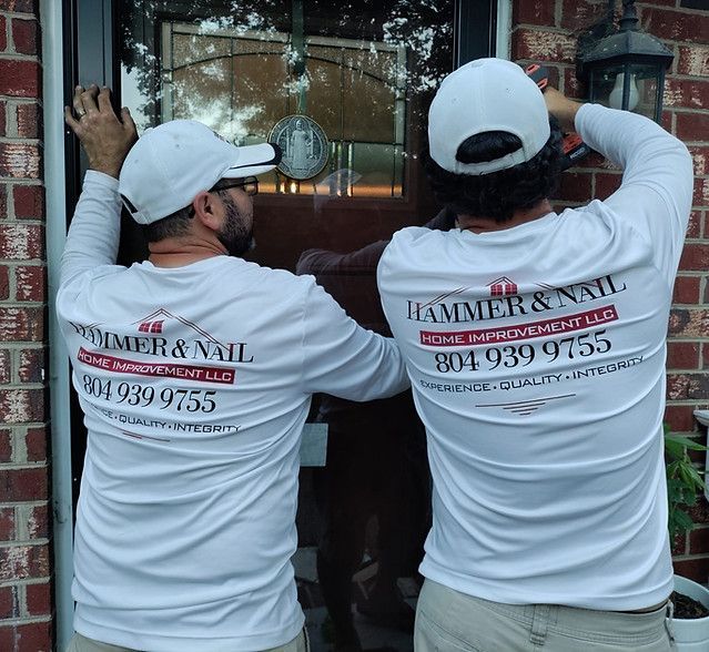 Two workers in white shirts and caps install a glass window at a brick building entrance.