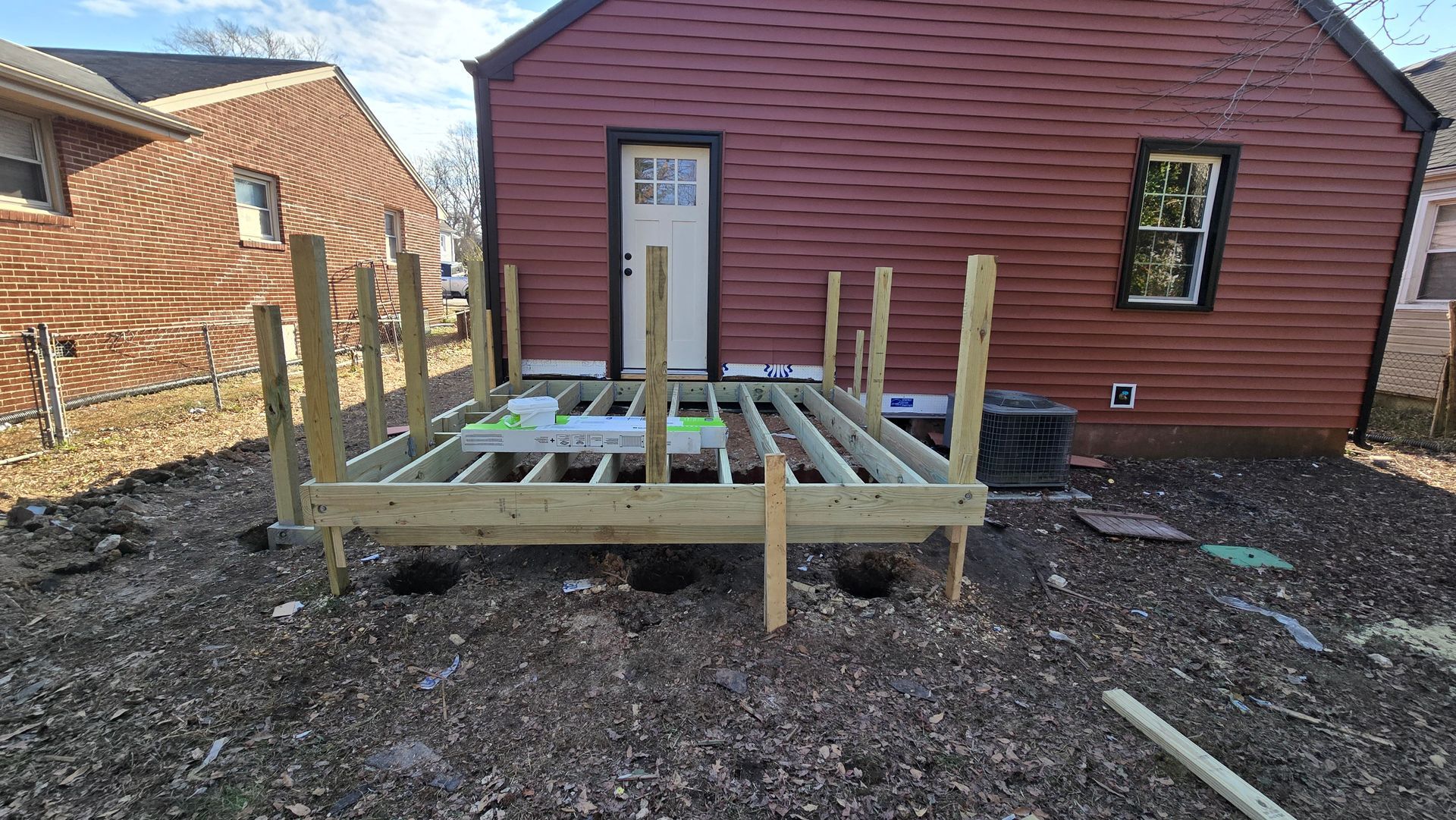Wooden deck frame under construction beside a red house, with upright posts and gravel yard.