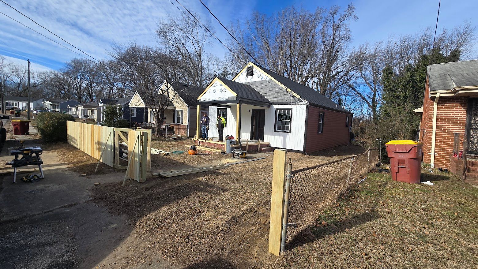 Small white house with red brick side wall, fenced yard, leafless trees, and a gravel driveway under a blue sky