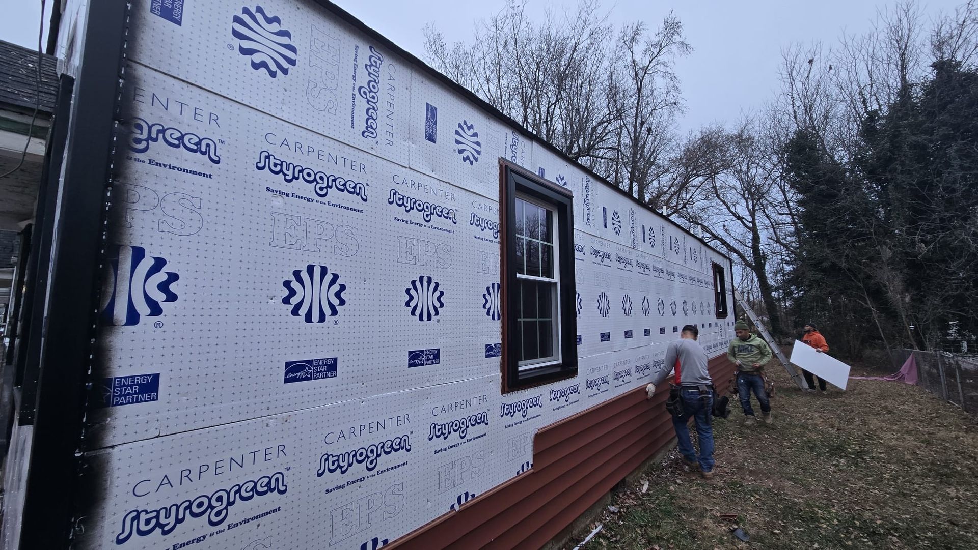 Two workers install white house wrap on a building exterior beside a wooded yard.