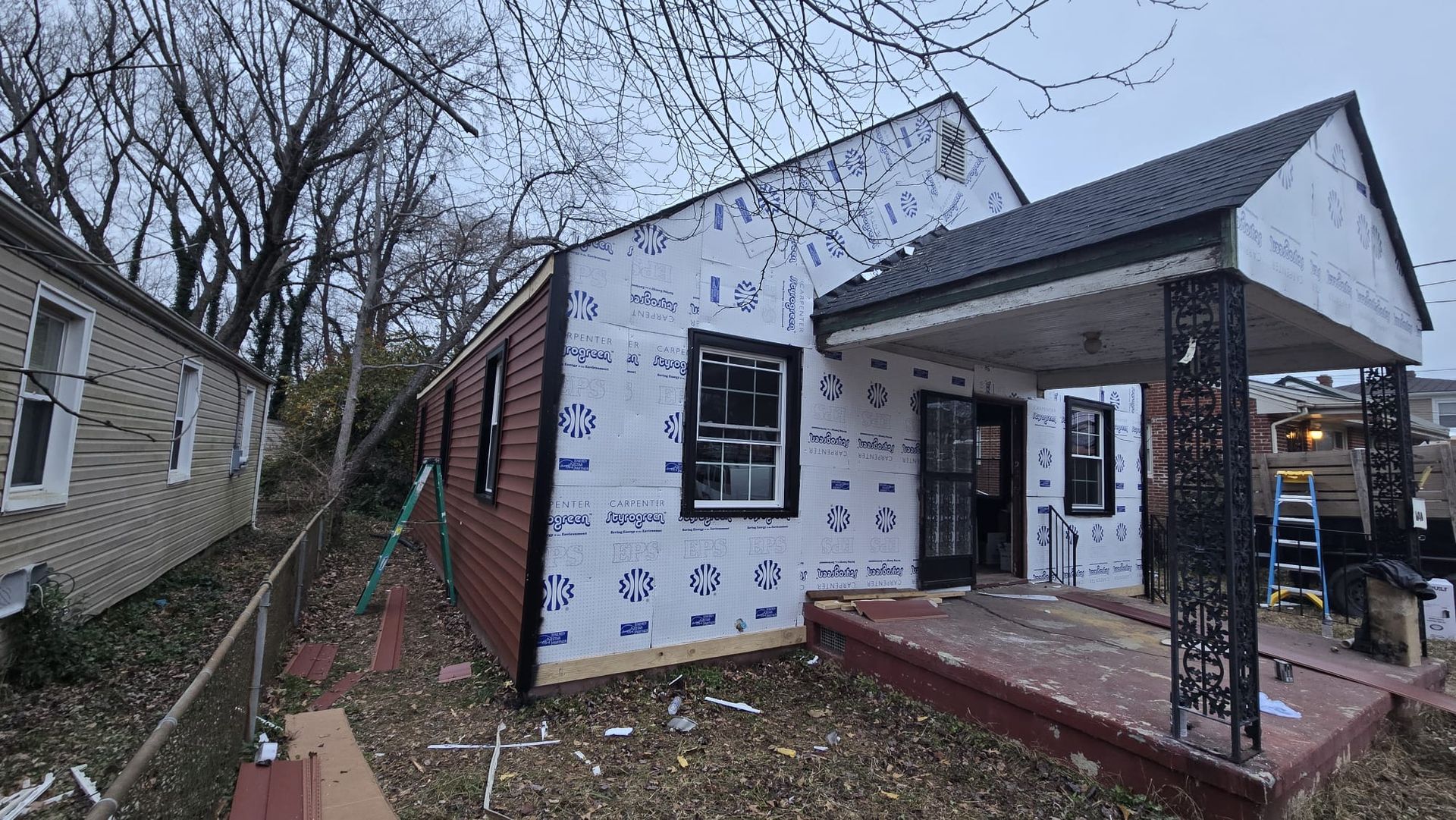 Partially renovated house exterior with white house wrap, black windows, and a covered front porch.