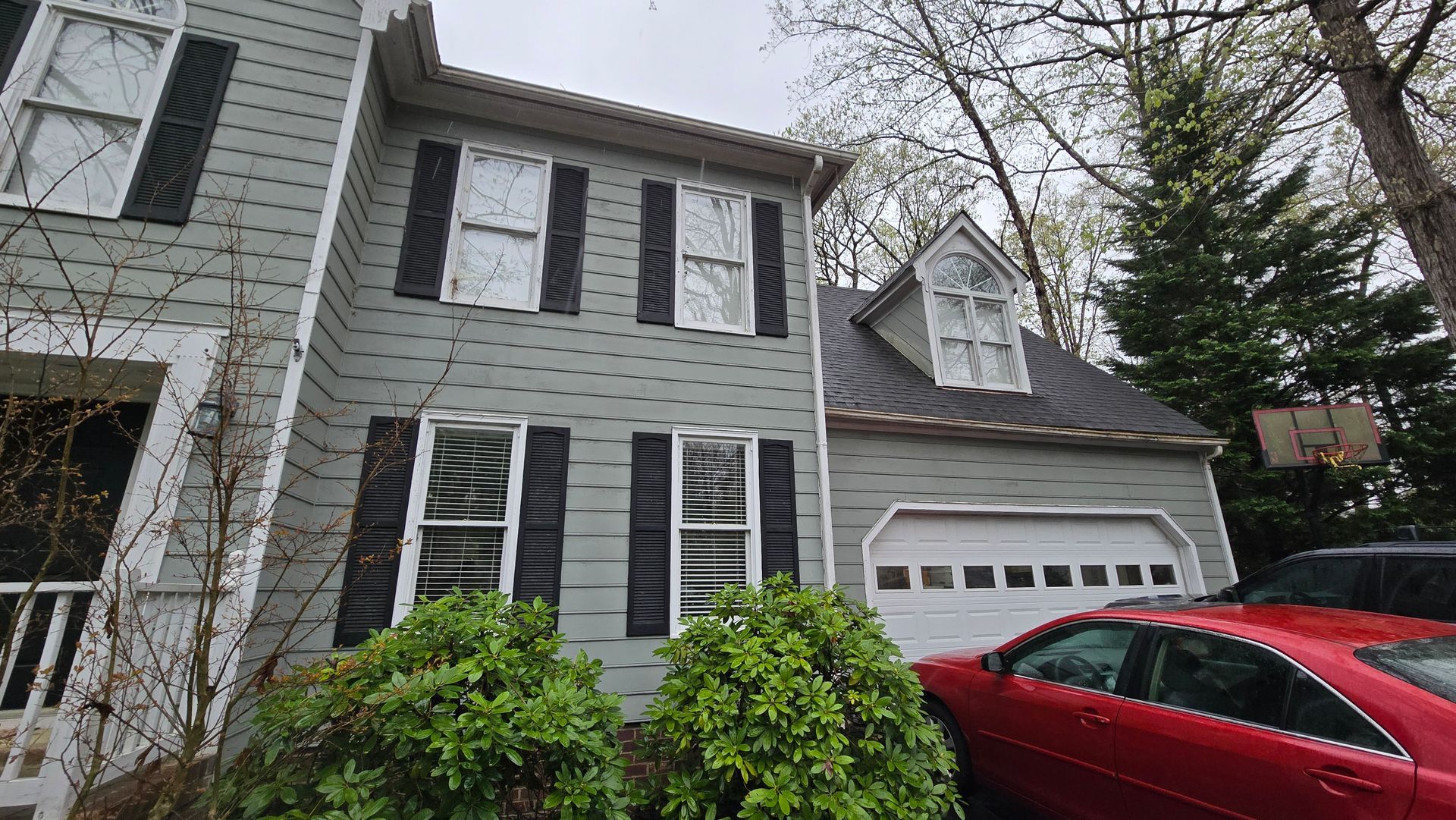 Gray two-story house with black shutters and attached garage, partially obscured by a red car and shrubs