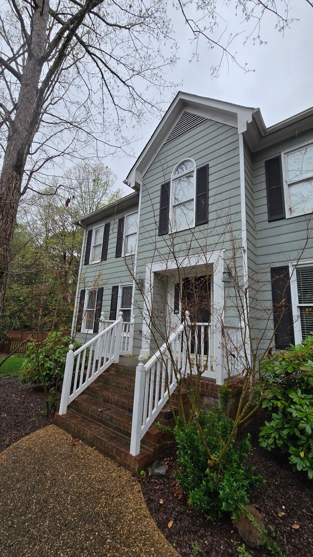 White two-story house with black shutters, front porch, and stairs on a rainy wooded hillside