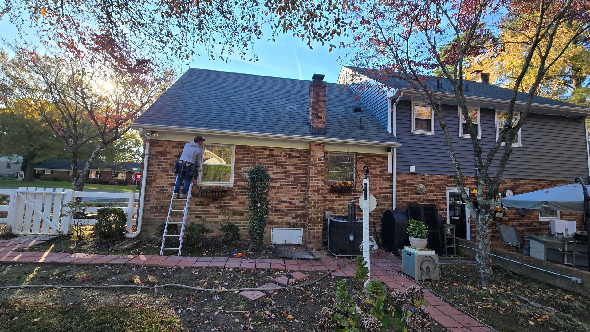 Worker on ladder repainting a brick house exterior in autumn with trees and yard visible