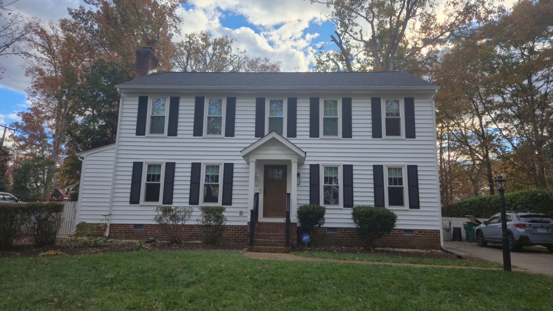 White two-story house with black shutters and a small front porch, viewed from the street under a partly cloudy sky