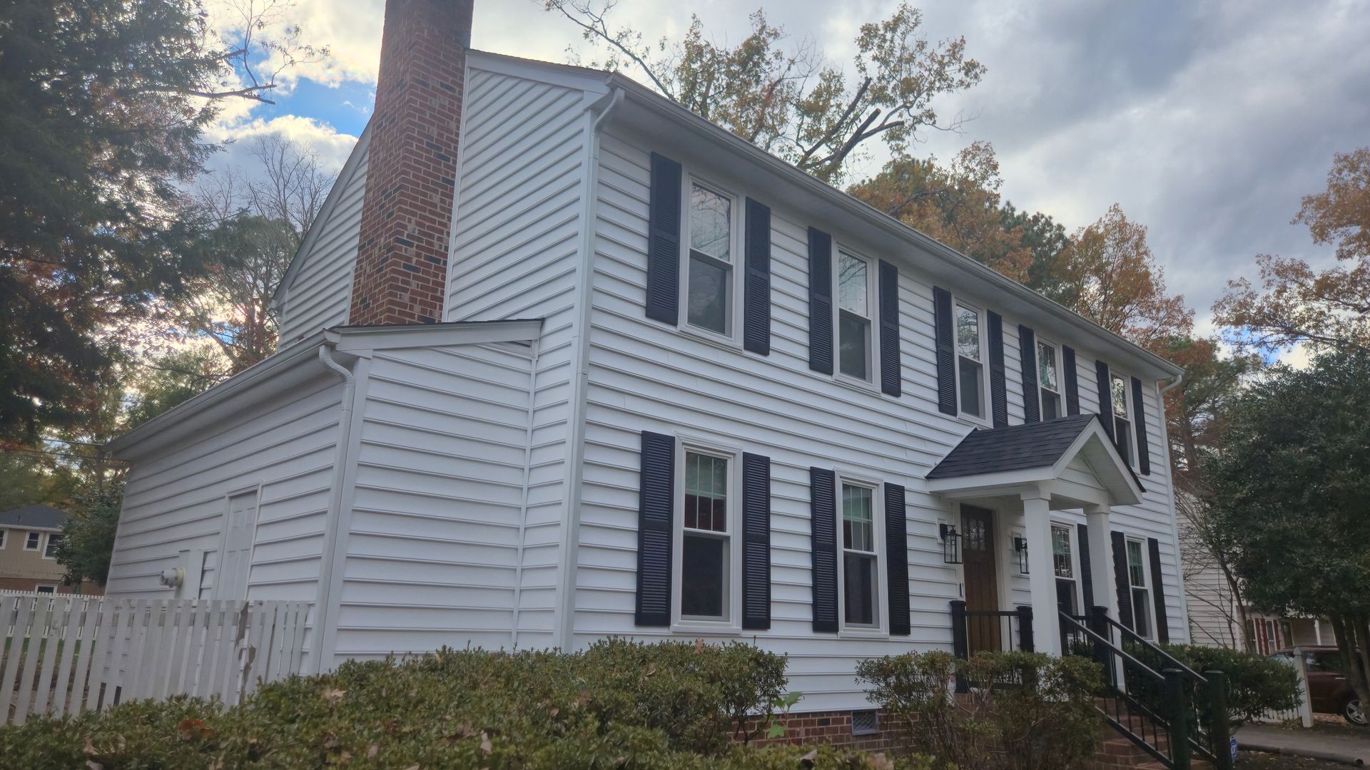 White two-story house with black shutters, brick chimney, and a front porch surrounded by trees.