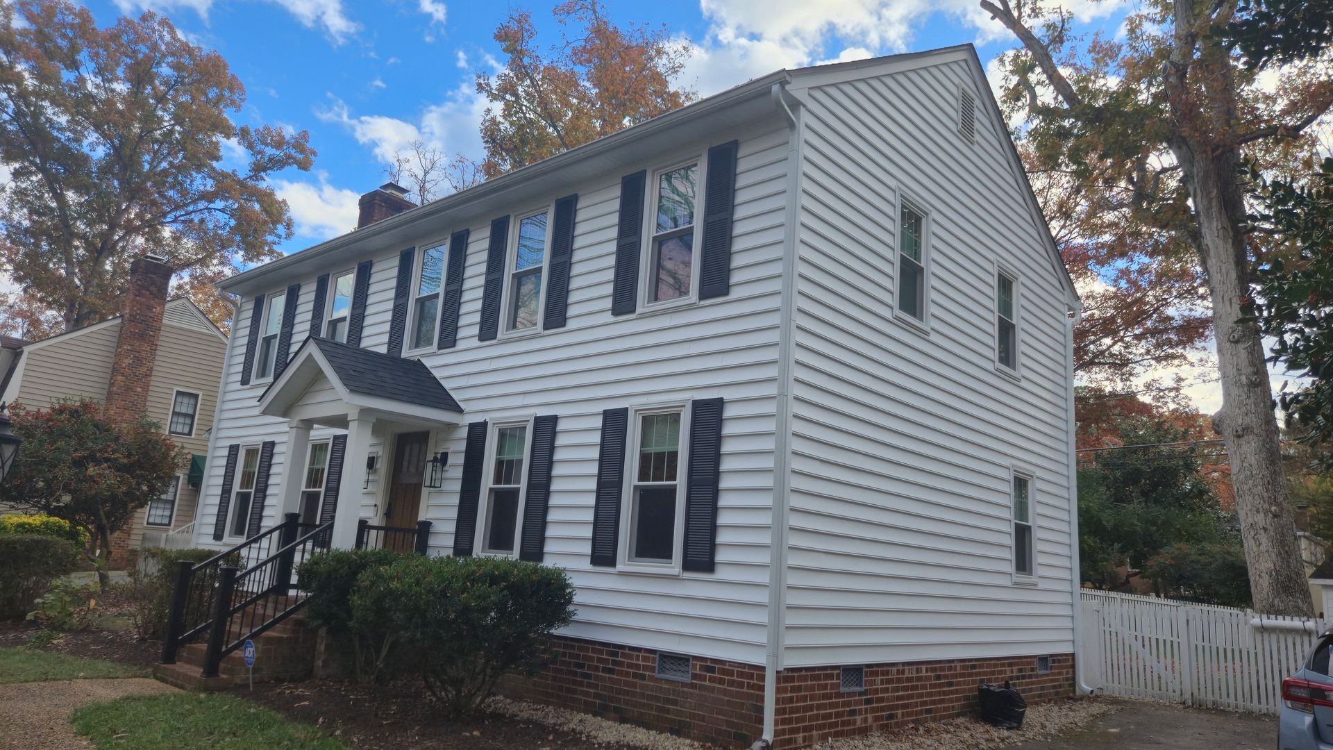 Two-story white house with black shutters, brick foundation, and autumn trees in the yard.