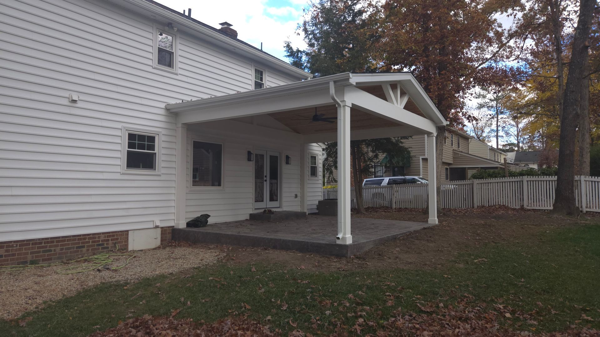 White house with a covered backyard patio and wooden deck, surrounded by autumn leaves and a fence.