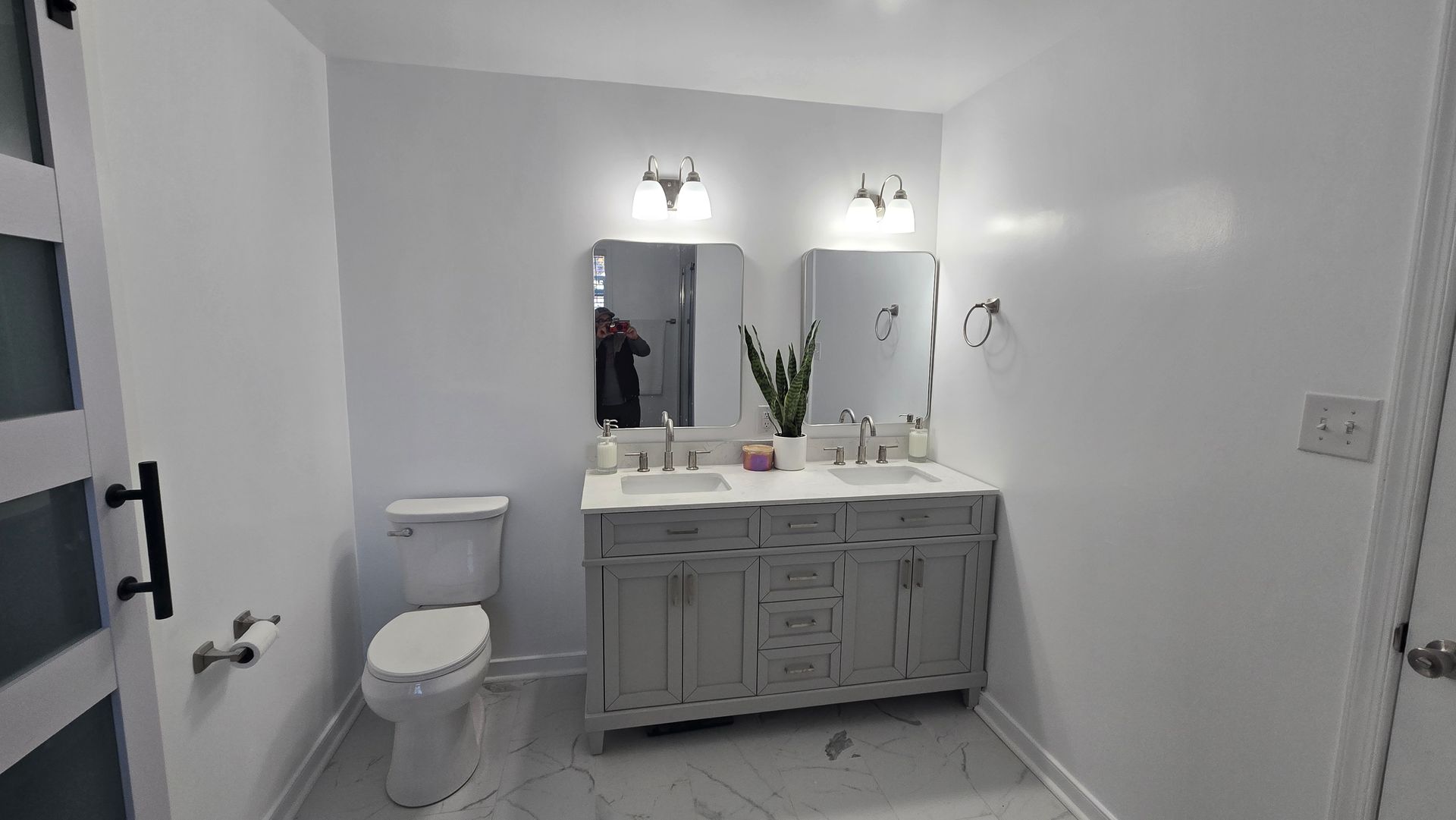 Small white bathroom with a toilet, double sink vanity, mirrors, and wall sconces.