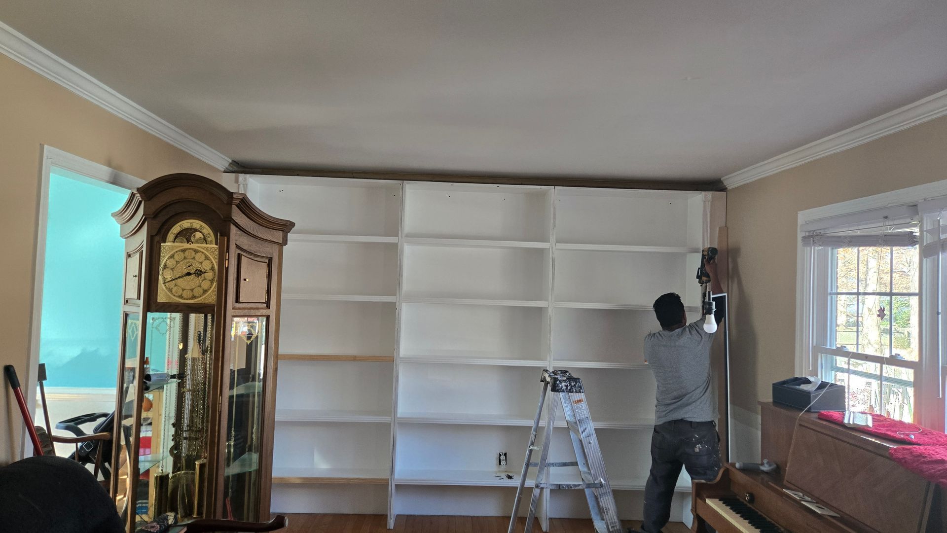 Person painting a tall white built-in bookshelf in a living room, with a ladder nearby.