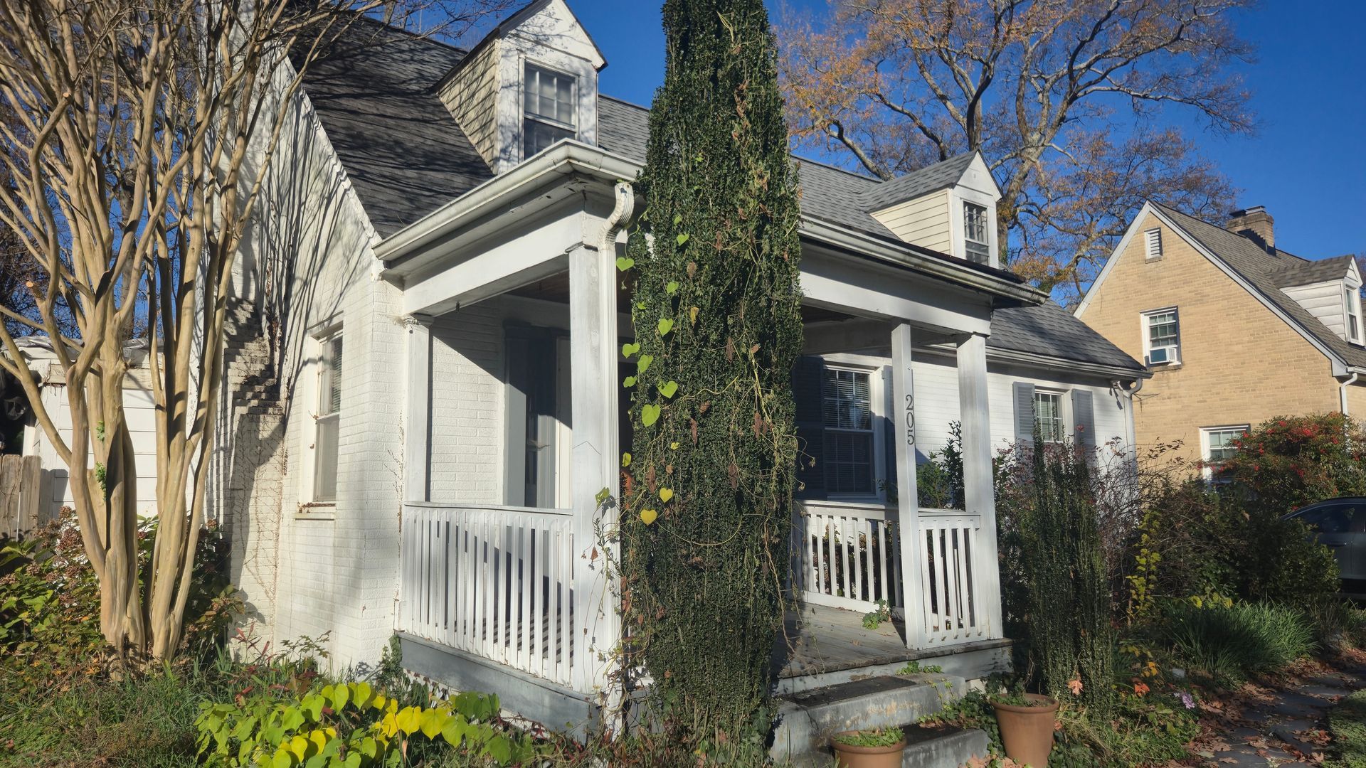 White cottage with a front porch and dormer windows, framed by tall trees and greenery in sunlight