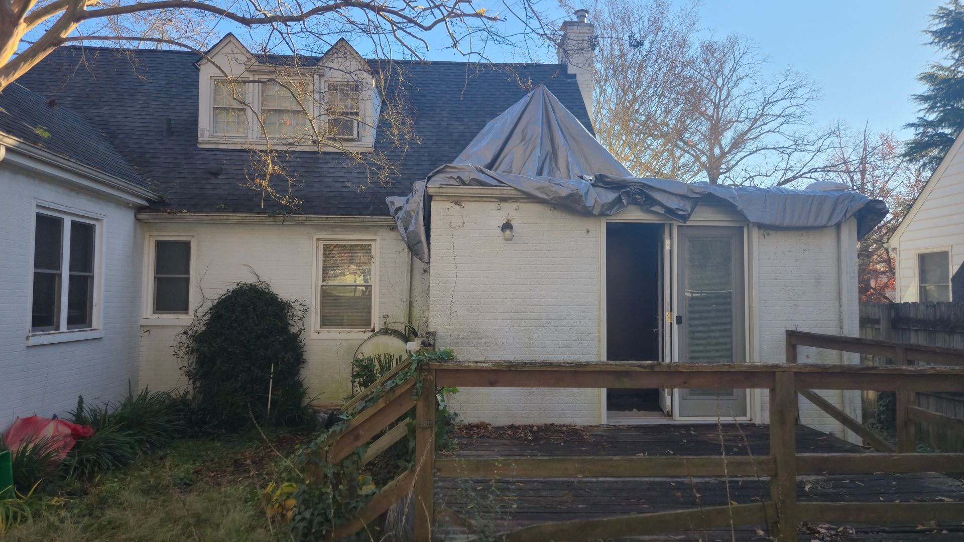 Damaged house exterior with boarded window, broken roof, and a wooden fence in front