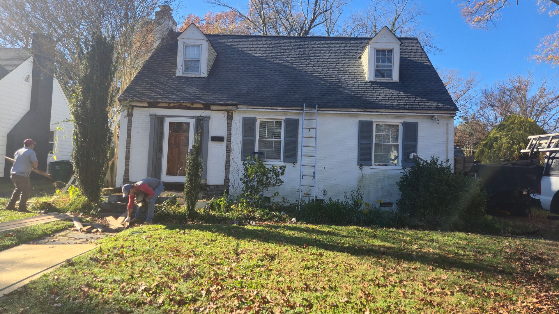 White house with a dark roof and dormer windows, front lawn, and a person near the left side.