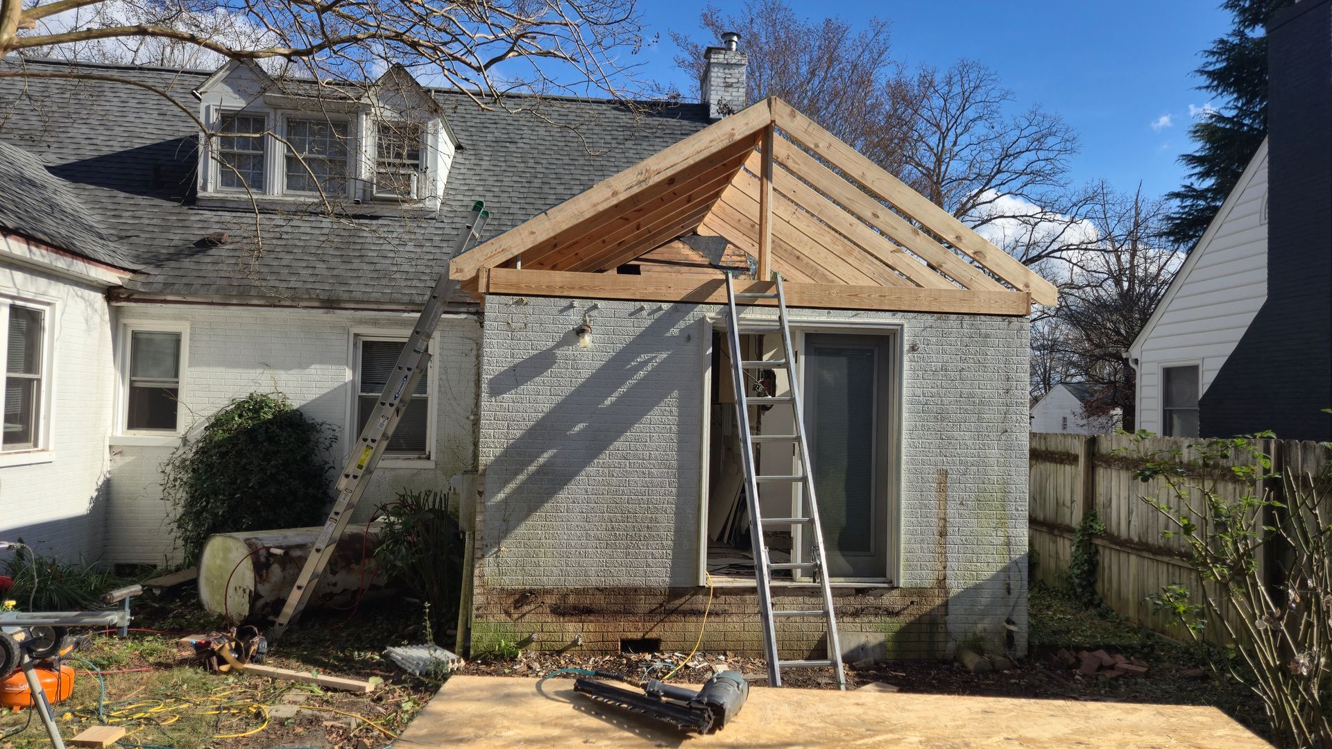 Small brick house with a partially built wooden porch roof; ladder and tools in a backyard.