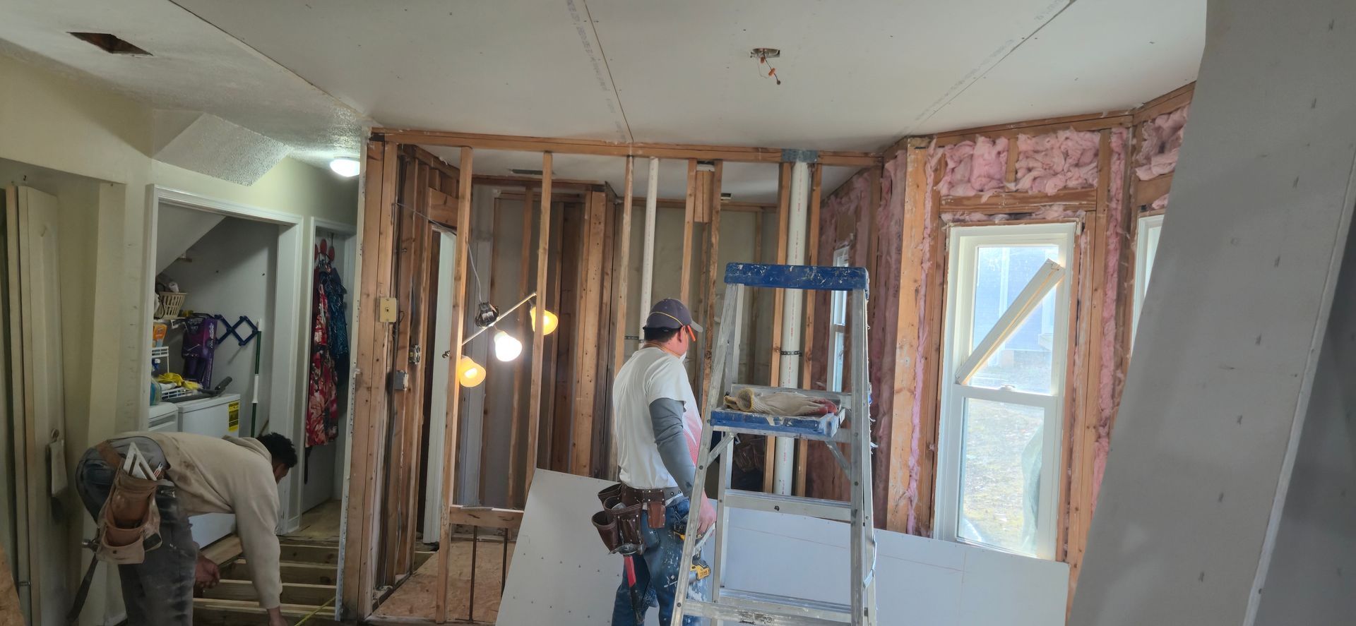 Person working in an unfinished room with exposed studs, drywall, and a ladder.