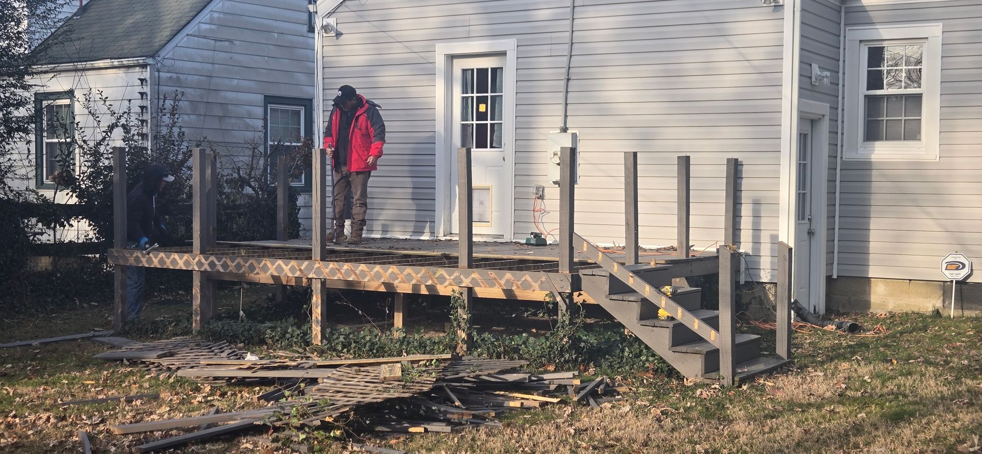 Person in a red jacket standing on a wooden deck beside a light gray house with steps and railings.