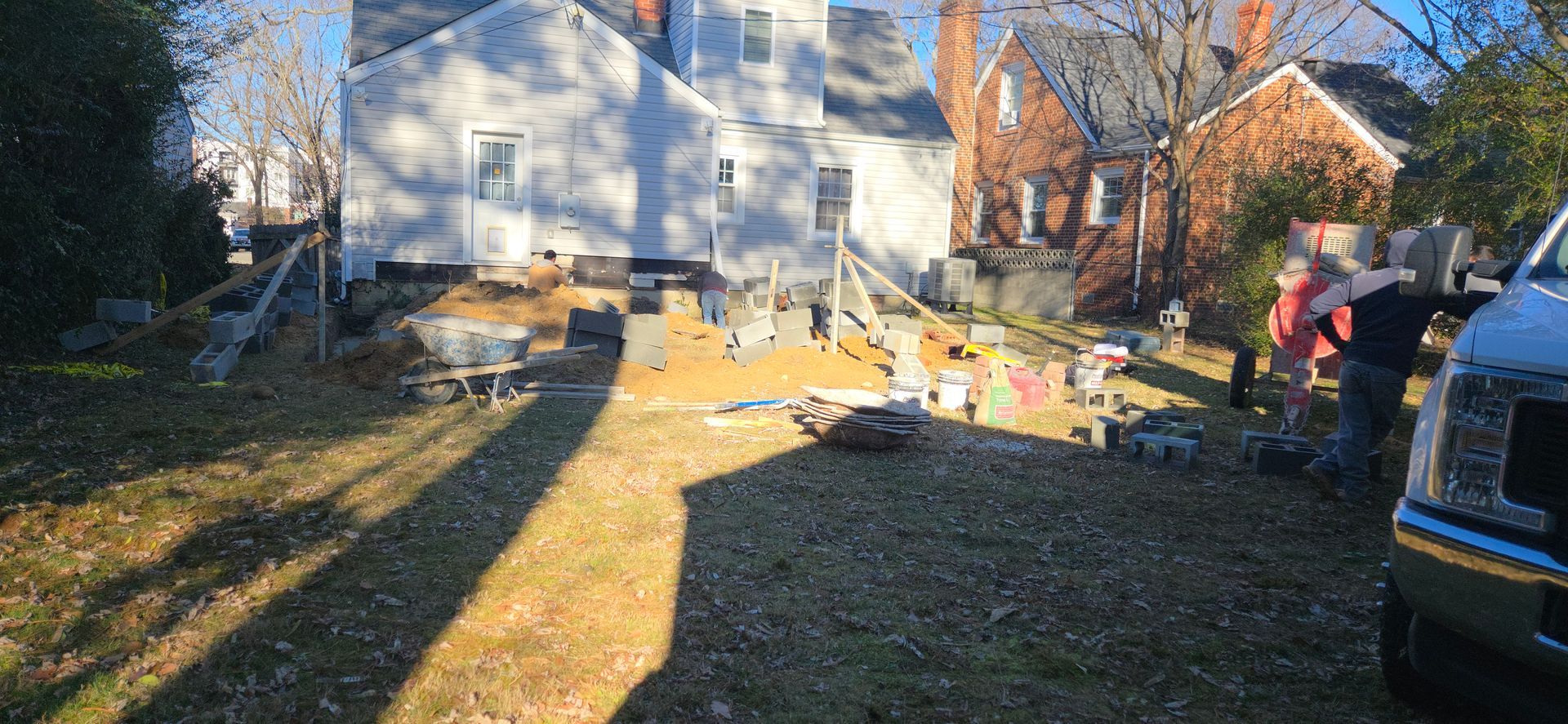 Sunlit backyard with leaf-covered grass, people near a house, and a parked SUV beside a fence