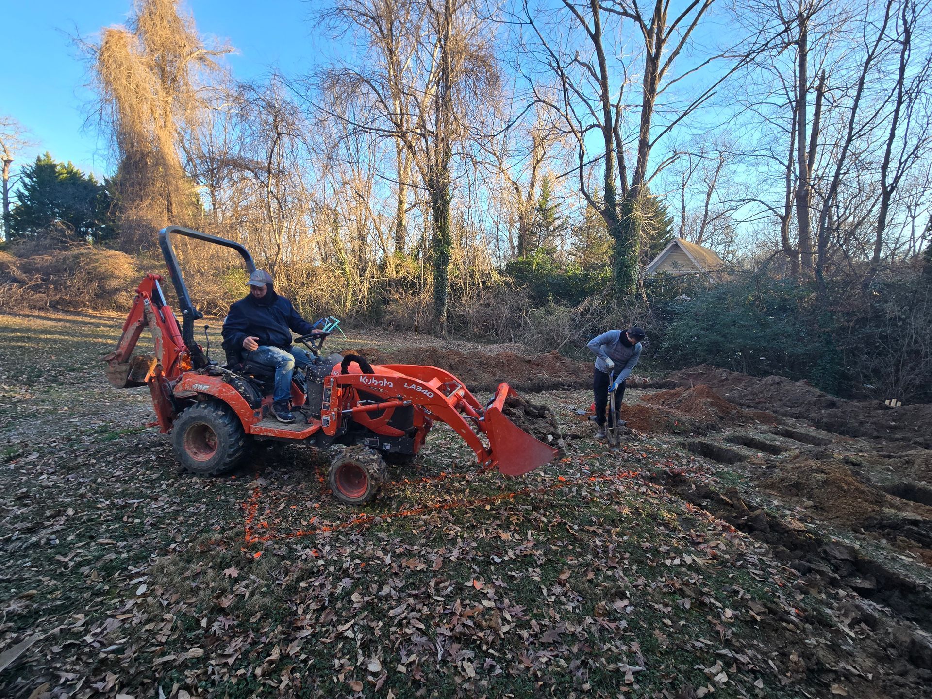 Red tractor with front loader moving leaves in a wooded yard, with two people nearby.