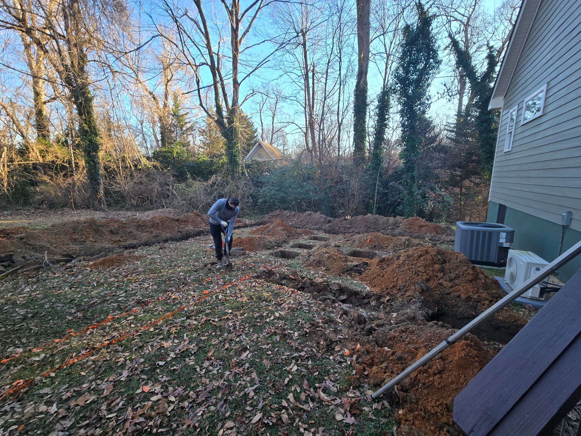 Person working in a leaf-covered backyard beside a house, with trees and a small pile of dirt.