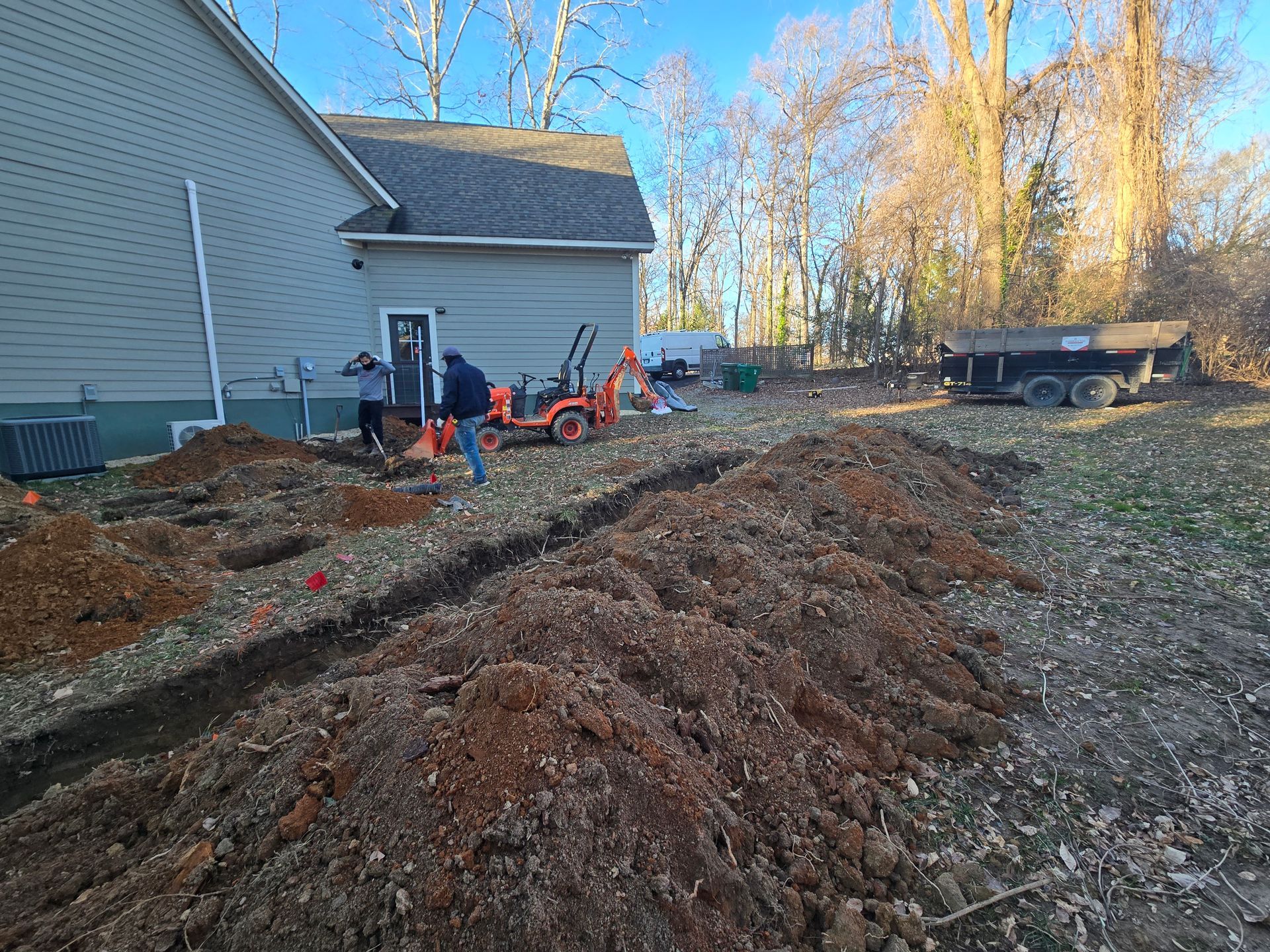 Construction equipment and workers grading a dirt trench beside a house in a backyard