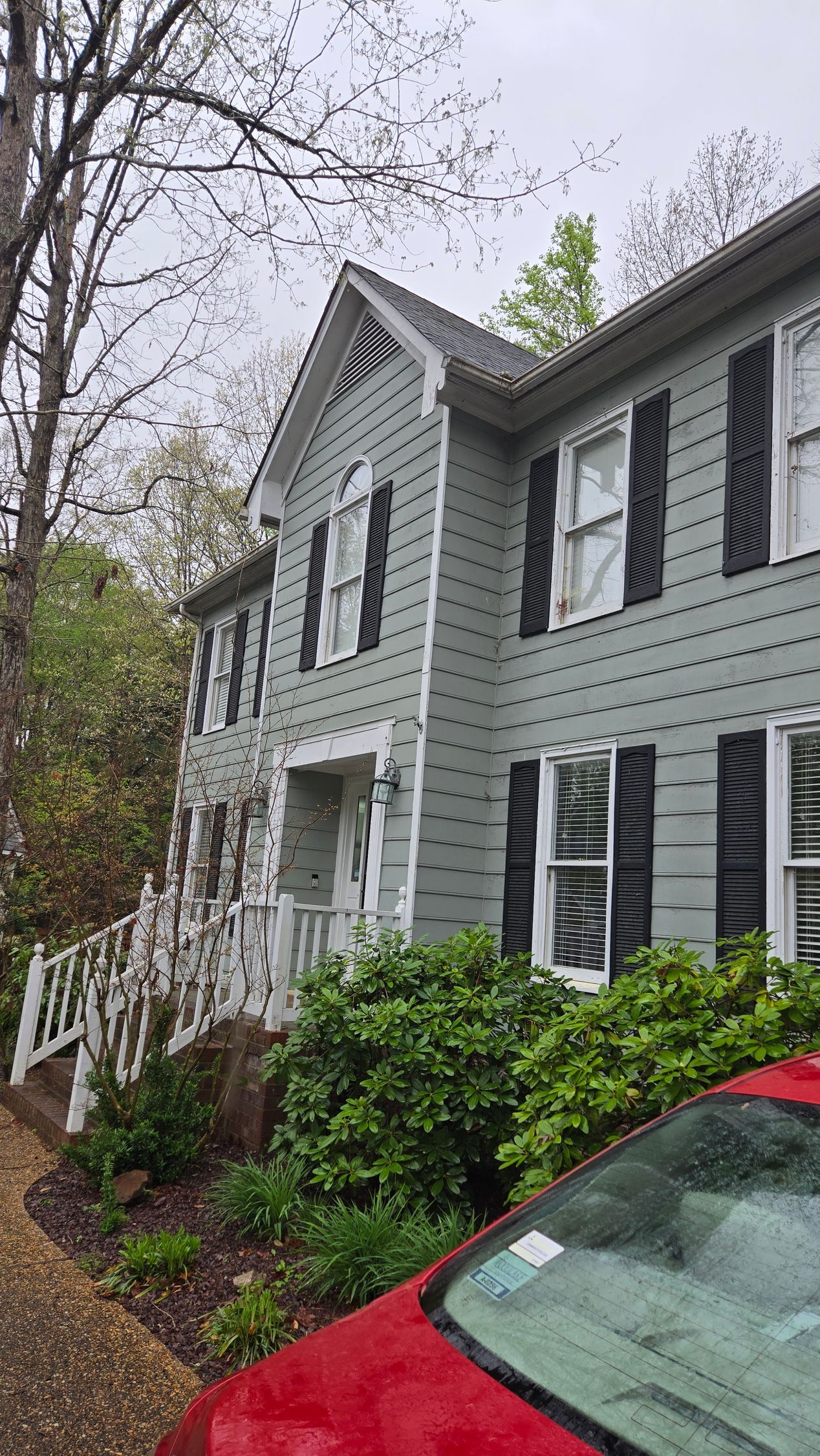Gray two-story house with white trim, black shutters, and a red car in front on a rainy day