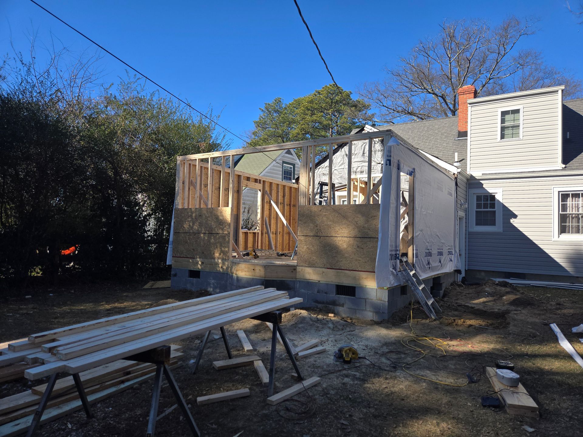 Partially framed house addition beside a white home, with lumber and sawhorses in the foreground.