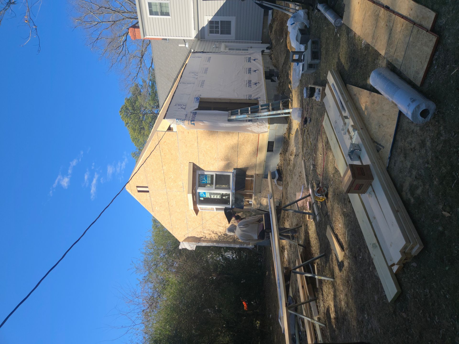 Backyard with a small beige house, bare trees, and scattered lumber and pipes under a blue sky