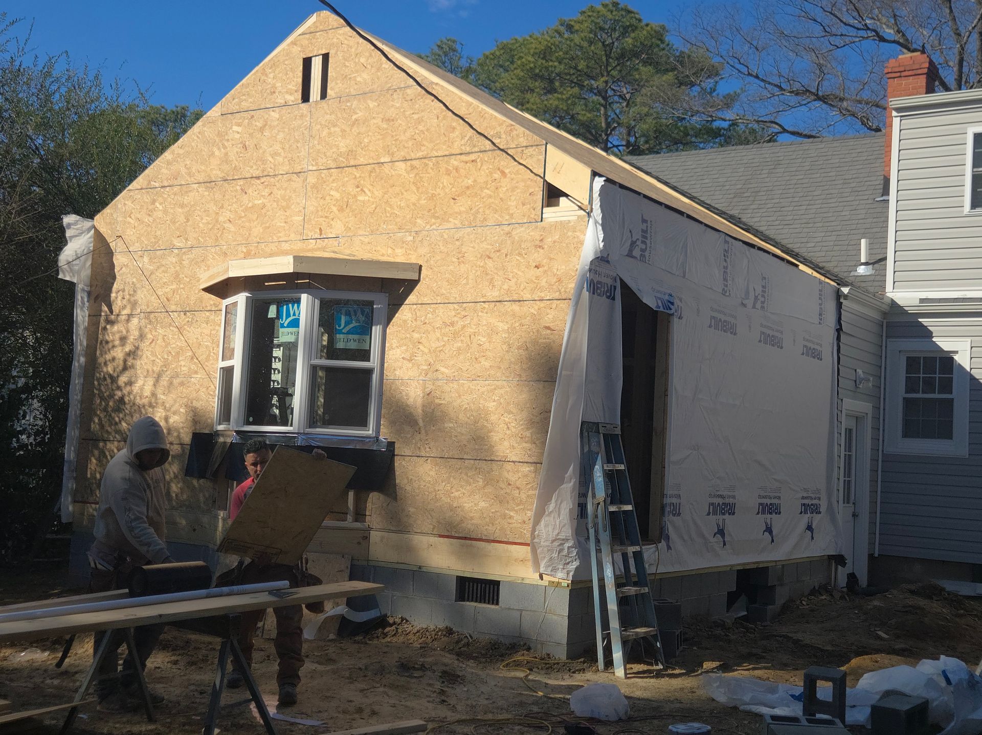 Small house under construction with exposed plywood walls, a window, and workers outside in daylight.