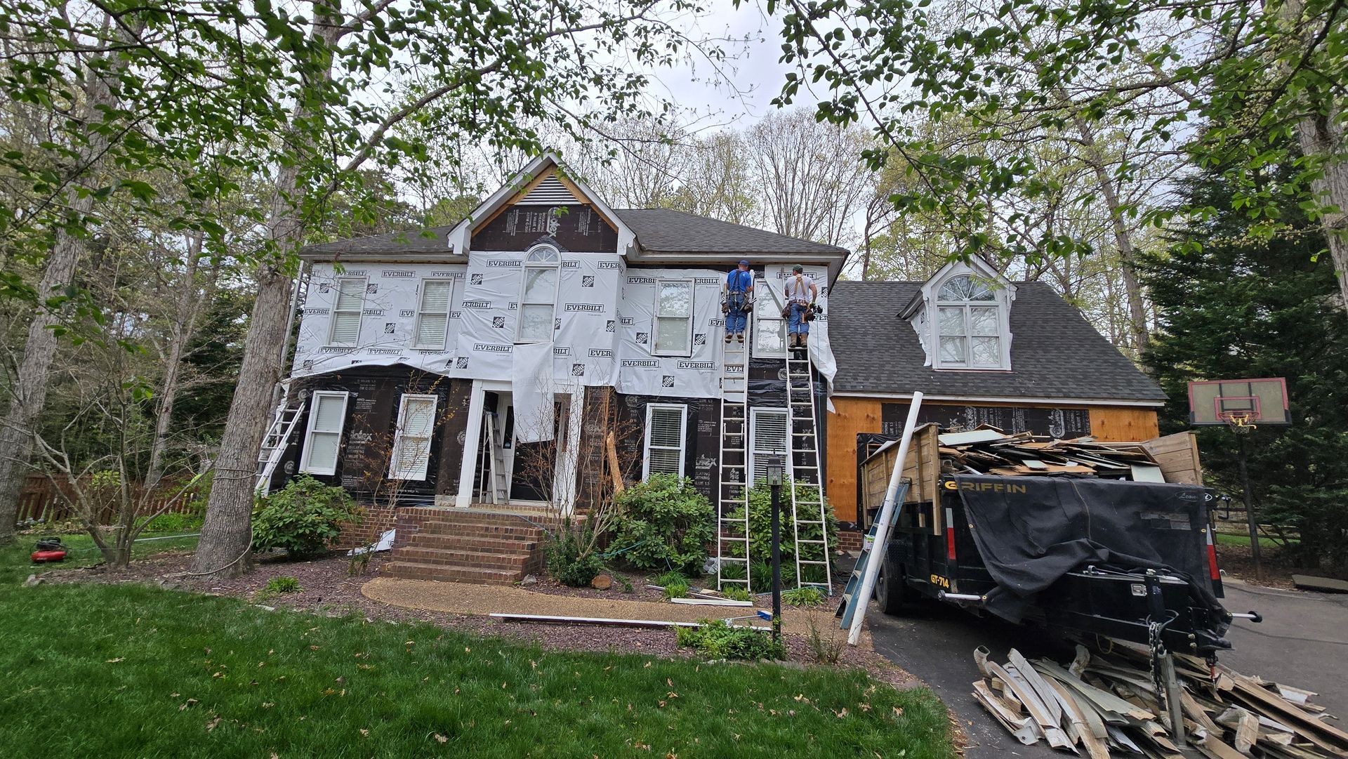 Two-story house under renovation with ladders, scaffolding, and construction materials in the driveway.